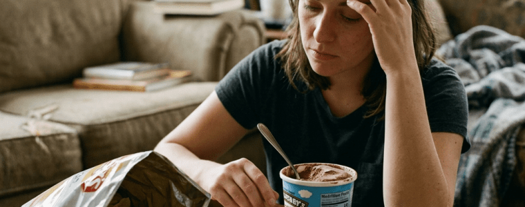 Woman sitting at a table with half-eaten pizza, chips, and a tub of ice cream, looking stressed